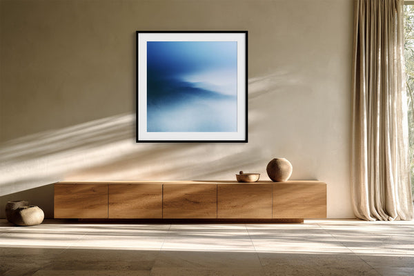 Black-framed blue abstract print above a long wooden sideboard in a sunlit space.