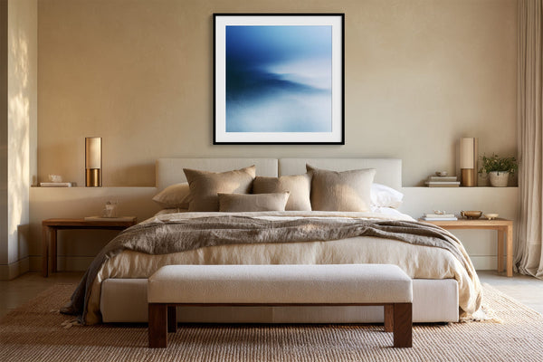 Black-framed blue abstract print centered above a bed with layered neutral bedding.