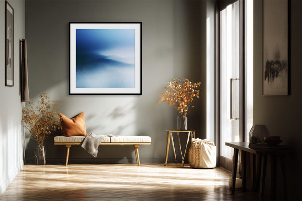 Black-framed blue abstract print above a bench with an orange cushion near tall windows.