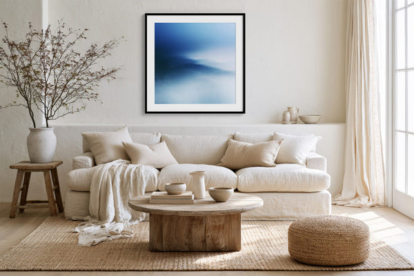 Black-framed blue abstract print above a white sofa, round wood table, and woven ottoman.