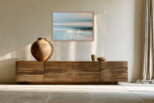 Light wood-framed coastal abstract hangs above a long wooden sideboard with ceramic vessels.