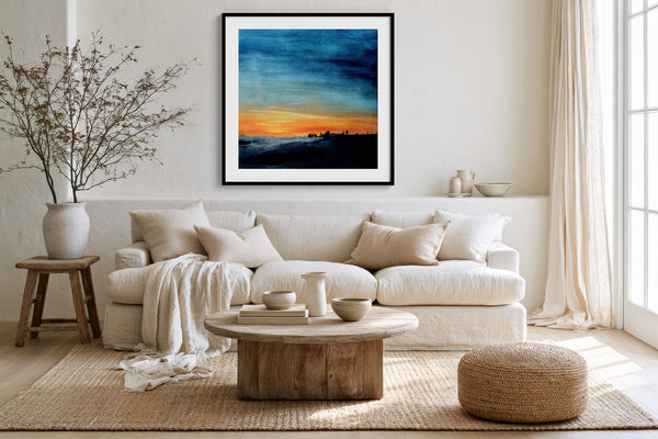 Black-framed abstract sunset print above a white sofa with pillows and a round wood coffee table.