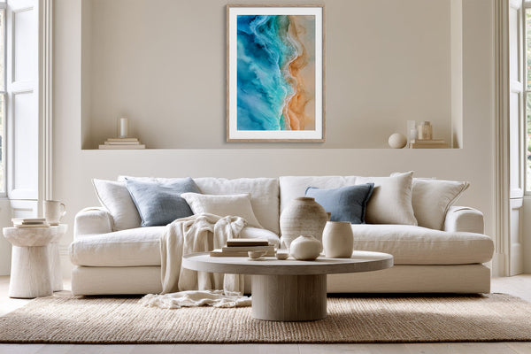 Framed coastal aerial artwork centered above a white sofa with blue-gray cushions.