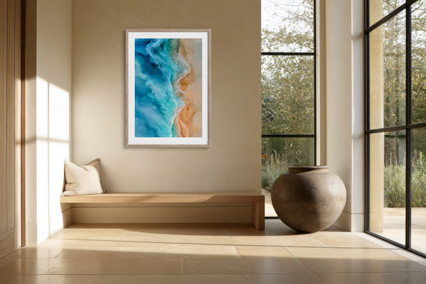 Framed turquoise-and-sand aerial artwork above a built-in bench beside large windows and ceramic vessel.