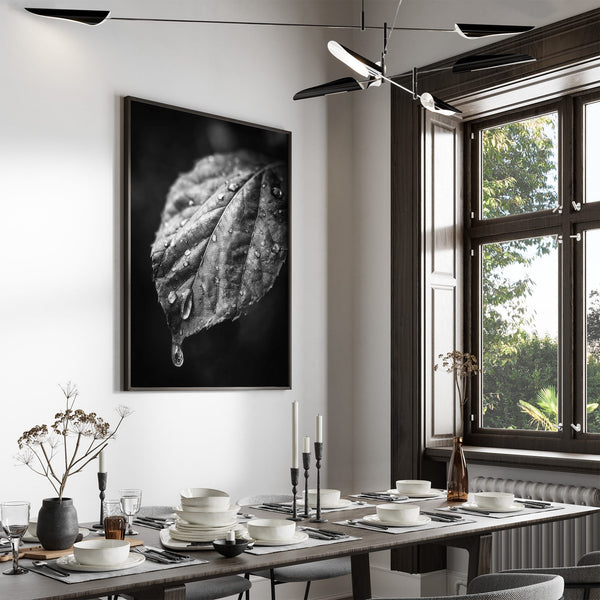 Large black-framed wet leaf photograph above a set dining table beside tall windows.