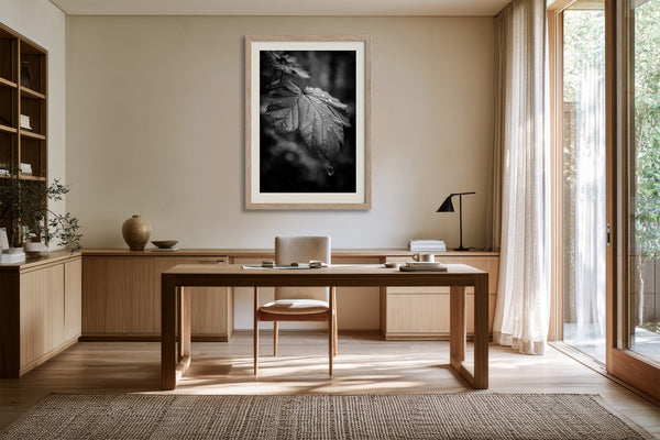 Framed black-and-white rain-covered leaf photograph above a wooden desk near tall glass doors.