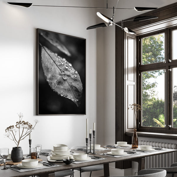 Framed black-and-white dew-covered leaf wall art displayed above a set dining table near tall windows.