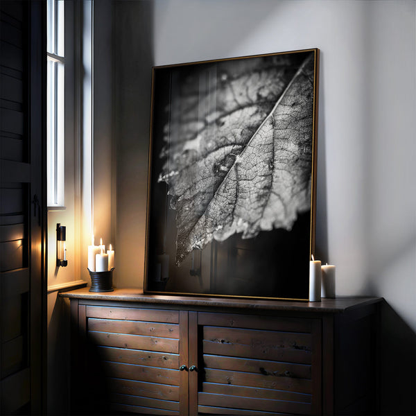 Glossy black-and-white leaf print in a thin gold frame resting on a dark cabinet.