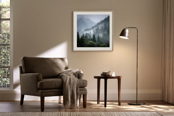 Framed misty mountain forest artwork above a brown armchair beside a floor lamp and side table.