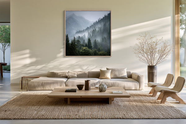 Misty mountain forest artwork in a slim wood frame above a low cream sofa and coffee table.