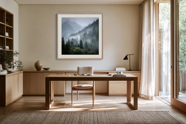 White-framed misty mountain forest artwork above a wooden desk with chair and black task lamp.