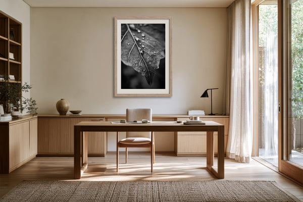 Framed monochrome leaf photograph displayed above a wooden desk beside tall glass doors.