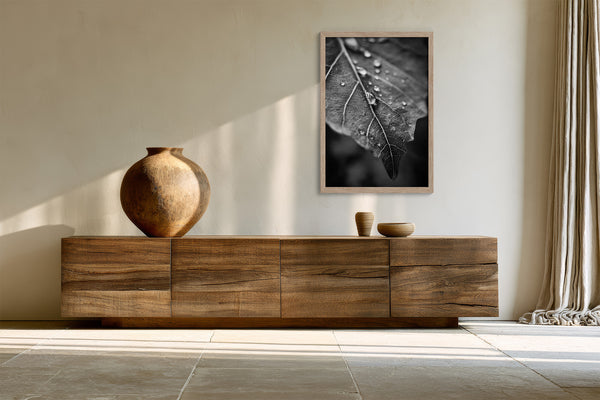 Framed monochrome leaf photograph above a low wooden sideboard with ceramic vase and bowls.