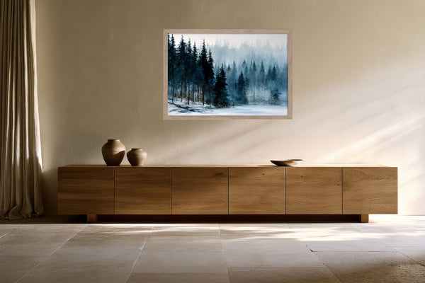 Framed winter pine forest artwork above a low wooden sideboard with ceramic vessels.