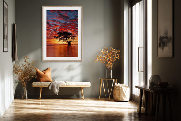 Light wood-framed sunset tree print above a bench beside tall windows.