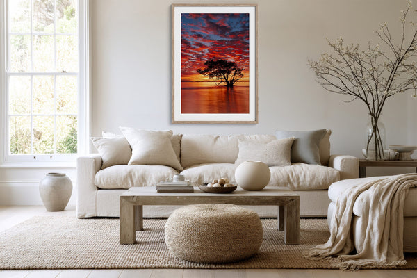 Light wood-framed sunset tree print above a white sofa, coffee table, and textured ottoman.