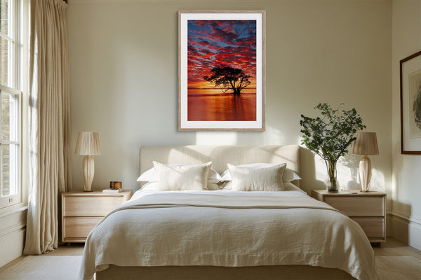 Light wood-framed sunset tree print centered above a neatly made bed.
