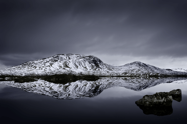 Snow-covered mountain ridge reflected in a still dark loch beneath a cloudy grey sky