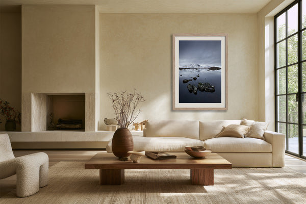 Light wood-framed mountain lake artwork above a cream sofa near a fireplace.