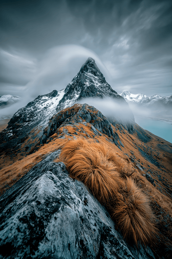 Snow-streaked mountain peak above a rocky ridge with ochre grasses and low cloud