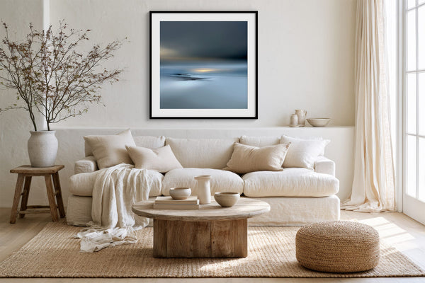 Black-framed Low Tide Light coastal print above a white sofa, wood coffee table, and woven ottoman.