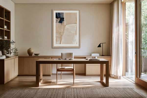 Framed beige and charcoal abstract artwork above a wooden desk beside tall windows.