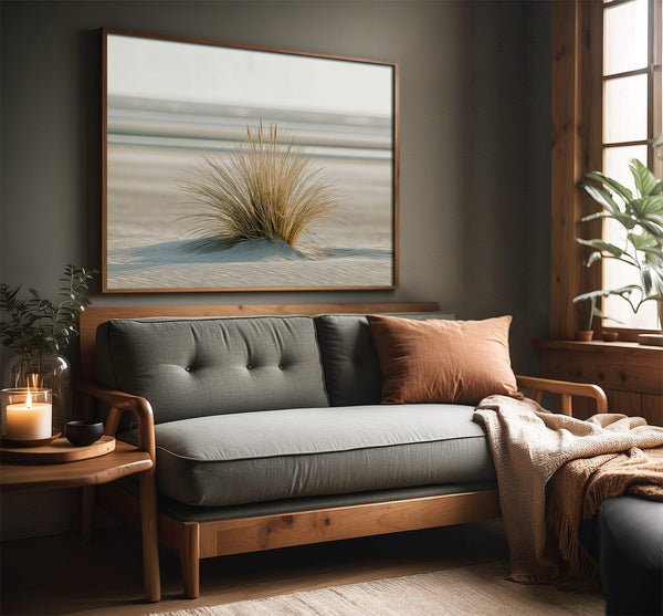 Dune grass coastal artwork in a dark wood frame above a gray sofa by a window.