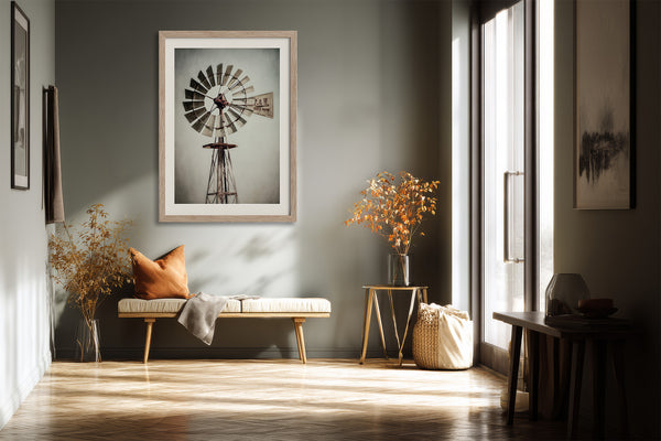 Light wood-framed windmill art above a bench with an orange pillow in a sunlit corridor.