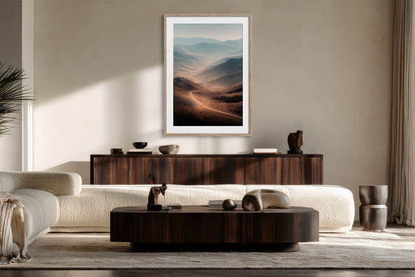Light wood-framed mountain valley artwork above a dark wood sideboard in a sunlit seating area.
