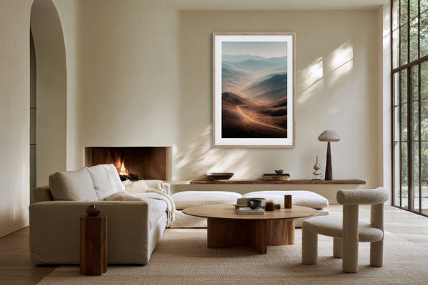 Light wood-framed mountain valley artwork above a fireplace and cream seating near grid windows.
