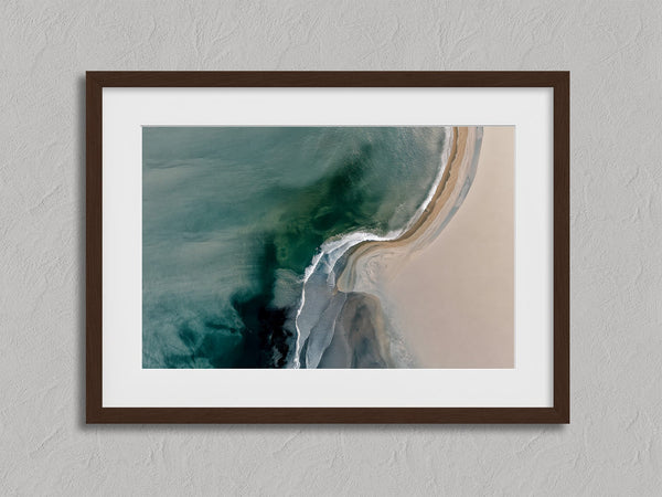 Aerial coastal view of deep teal water curling into pale sand along a sweeping shoreline.