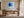 Black-framed blue abstract print above a wooden desk beside tall windows.
