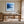 Black-framed blue abstract print above a wooden desk beside tall windows.