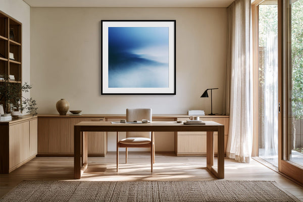 Black-framed blue abstract print above a wooden desk beside tall windows.
