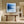 Black-framed blue abstract print above a rustic wooden bench in a bright hallway.