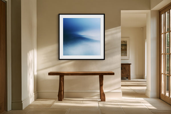 Black-framed blue abstract print above a rustic wooden bench in a bright hallway.