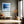 Black-framed blue abstract print above a bench with an orange cushion near tall windows.