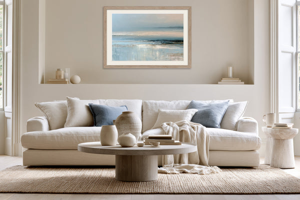 Light wood-framed coastal abstract above a white sofa with pale blue cushions.