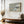 Framed Pale Estuary artwork above a wooden bench, with a vase of dried branches by tall windows.