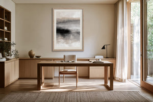 Framed abstract landscape in grey and charcoal above a wooden desk beside tall windows.