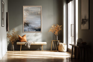 Framed grey and charcoal abstract artwork above a bench with an orange cushion.