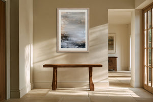 Framed layered abstract artwork hanging above a rustic wooden bench in a sunlit hall.