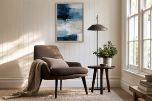 Framed blue and white abstract artwork above a brown armchair beside a floor lamp and window.
