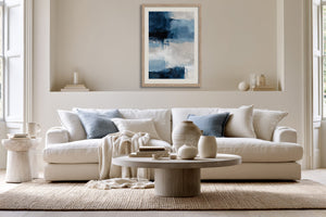 Framed blue and white abstract artwork centered above a white sofa with pale blue pillows.