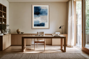 Framed blue and white abstract artwork above a wooden desk with chair and floor lamp.