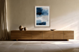 Framed blue and white abstract artwork above a long wooden sideboard with ceramic vessels.