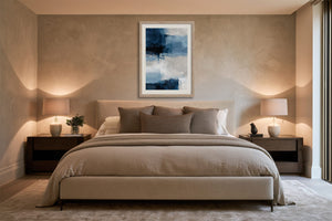 Vertical blue and white abstract artwork in a light wood frame above a bed with neutral bedding.