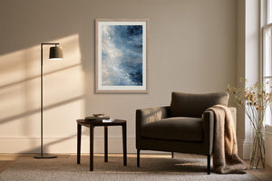 Blue and white abstract wall art in a light wood frame above an armchair and side table.