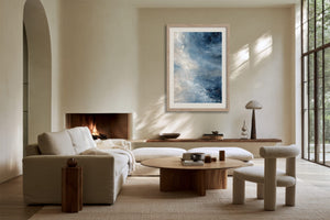 Blue abstract wall art in a pale wood frame above a fireplace in a sunlit sitting area.