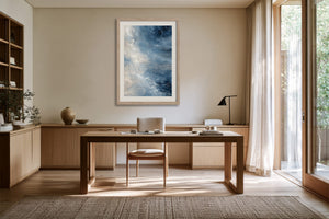 Framed blue abstract artwork hanging above a wooden desk beside floor-to-ceiling windows.